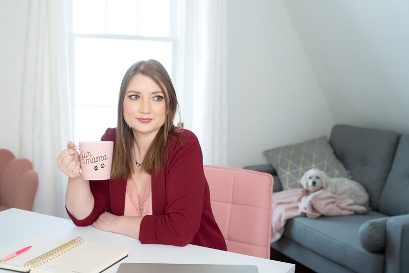 Tara Reid at her desk holding a coffee mug, with her small white rescue dog on the couch behind her